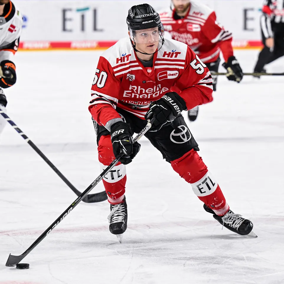 A hockey player in a red jersey with the number 50 and 'Rhein Energie' printed on it, playing on the ice and controlling a puck with his stick.